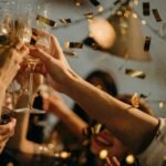 A lively indoor party scene with people toasting champagne glasses amid confetti and warm lights.