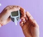 Hands holding a glucometer to test blood sugar levels on a purple background.