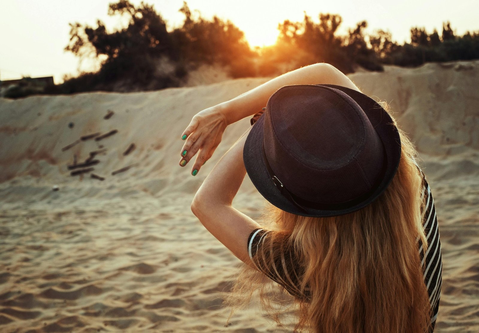 A woman wearing a fedora enjoys the serene sunrise at a sandy beach.